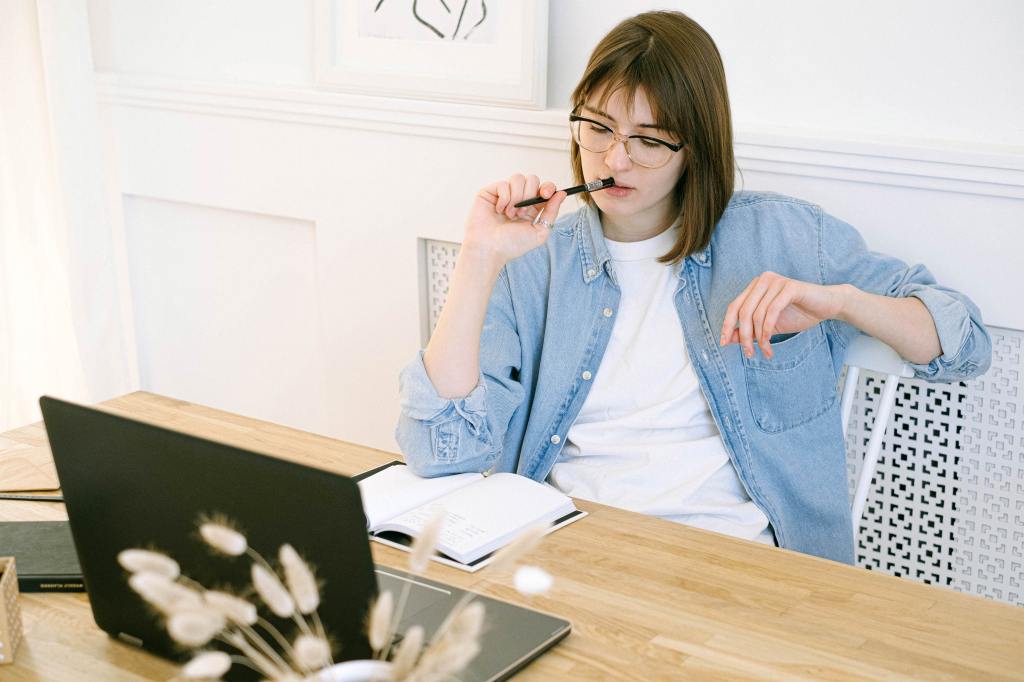Mujer pensativa está sentada frente a computadora sosteniendo un lapiz y mirando hacia un cuaderno que está también frente a ella.