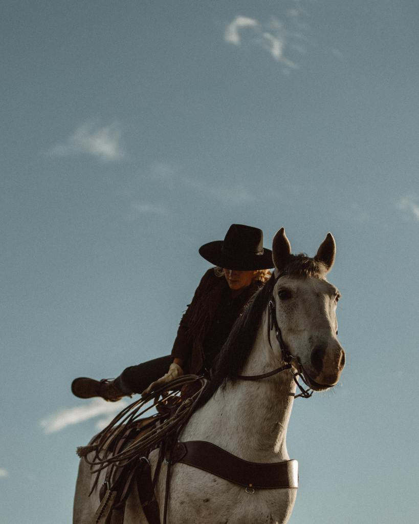 Una mujer con un sombrero de cowboy va subiéndose a un caballo blanco. 