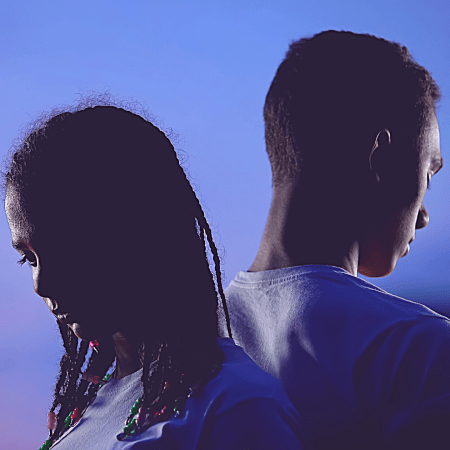 Una pareja de personas ven hacia el suelo. Los dos están apoyando su espalda en la del otro y están iluminados con una luz dramática.