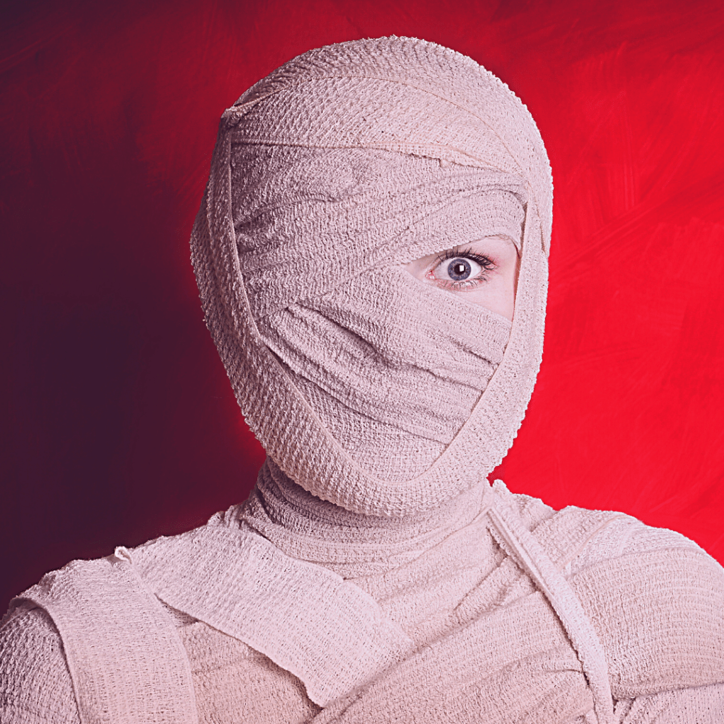 Una mujer vendada de todas partes del cuerpo, menos de un ojo que ve a la cámara. Simula ser una momia. El fondo de la imagen es rojo "sangre".