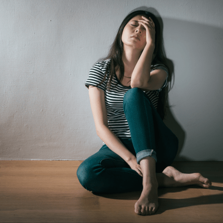 Mujer sentada en el piso tiene una mano reposando en su pierna y la otra tocándose la frente. Tiene los ojos cerrados con actitud de frustración y tristeza.