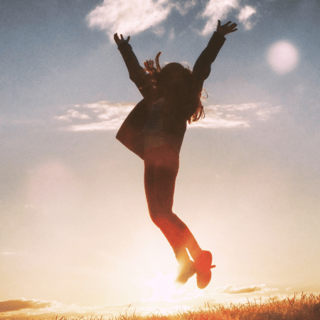 Una mujer, a contraluz, salta, con los brazos al aire, en señal de triunfo. Se encuentra al aire libre.