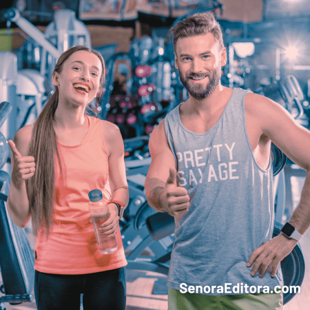 Una pareja, un hombre y una mujer están en un gimnasio, usan ropa deportiva y saludan a la cámara haciendo una señal con un pulgar hacia arriba.